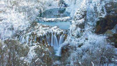 A frozen waterfall cascades over rocks into a lower body of water, surrounded by snow-covered trees and cliffs in a scenic winter landscape.